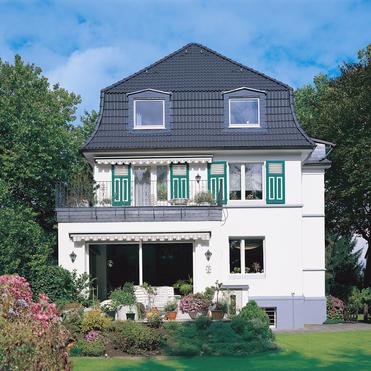 Helles, zweigeschossiges Einfamilienhaus mit grünen Fensterläden, Balkon und gepflegtem Garten unter blauem Himmel.
