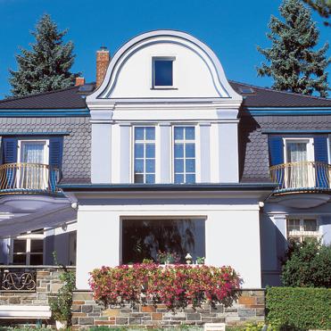 Fassade eines eleganten Hauses mit Erker, Balkonen und blühenden Pflanzen vor blauen Himmel und Bäumen.