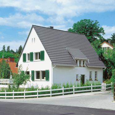 Weißes Wohnhaus mit grünem Fensterladen, umgeben von Bäumen und einem Garten, an einer Kurve in der Straße.