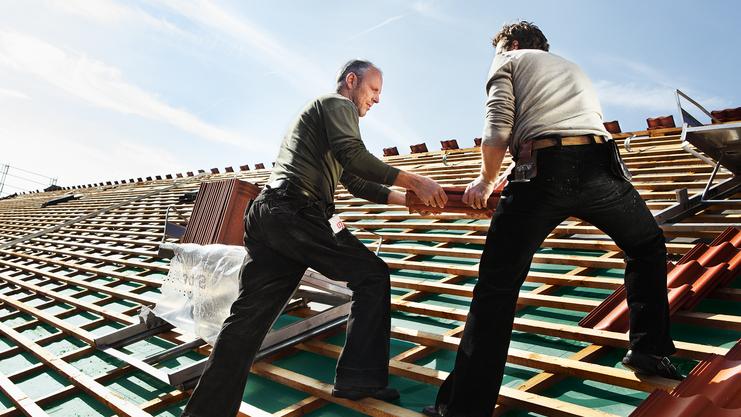 Zwei Handwerker arbeiten auf einem geneigten Dach, während sie Holzlatten tragen und das Dach decken.