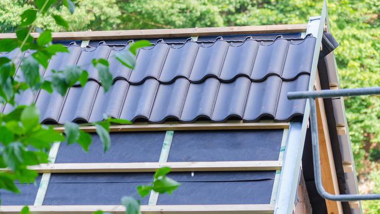 Dach mit schwarzen Dachziegeln und darunter liegender Unterkonstruktion, umgeben von grünen Blättern.