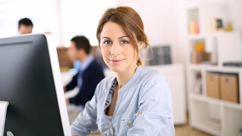 Junge Frau sitzt vor einem Computer in einem hellen Büro, lächelnd und aufmerksam, im Hintergrund Kollegen bei der Arbeit.