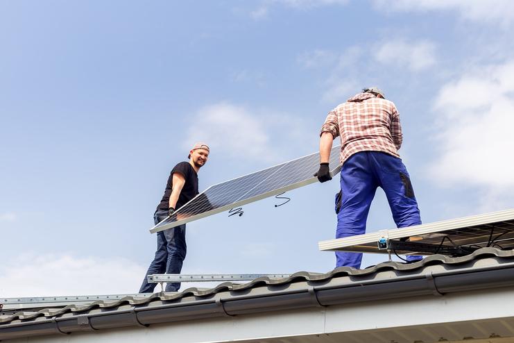 Zwei Männer installieren ein Solarpanel auf einem Dach unter blauem Himmel.