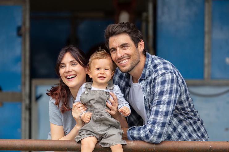 Lächelnde Familie, bestehend aus einem Mann, einer Frau und einem Kleinkind, posiert am Zaun eines Pferdestalls.