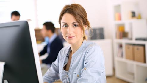 Junge Frau sitzt vor einem Computer in einem hellen Büro, lächelnd und aufmerksam, im Hintergrund Kollegen bei der Arbeit.