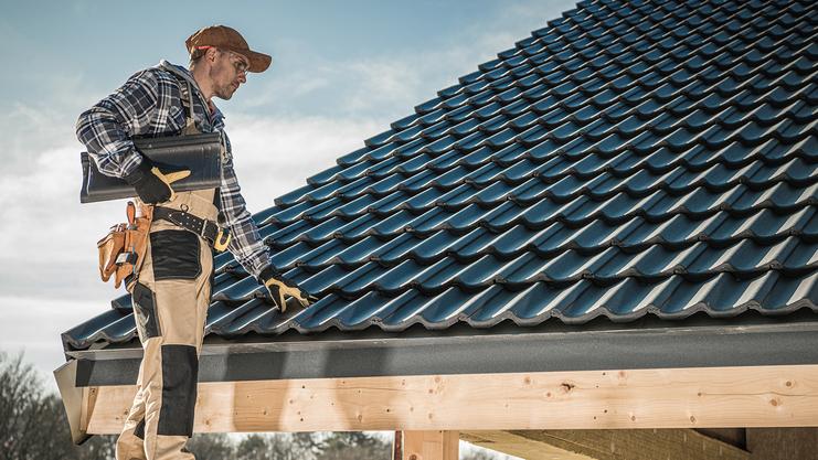 Dachdecker mit Unterlagen steht auf einem Dach mit blauen Ziegeln und prüft die Arbeitshöhe unter einem klaren Himmel.