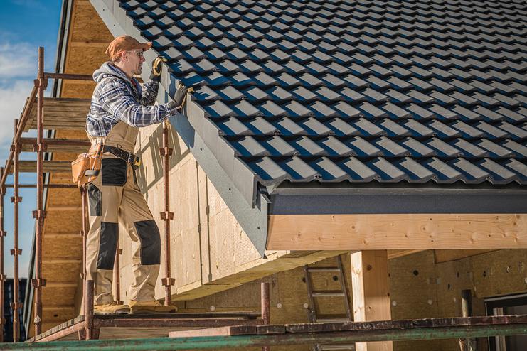 Handwerker auf einem Gerüst installiert Dachziegel an der oberen Kante eines Hauses unter klarem Himmel.