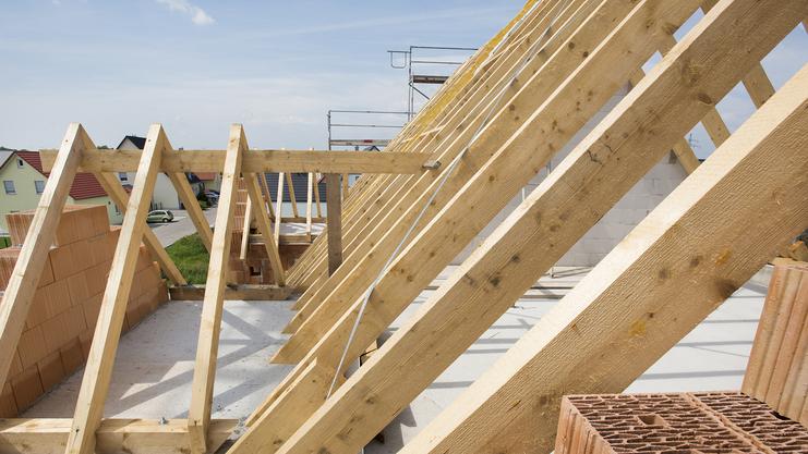 Blick auf eine Dachkonstruktion mit Holzsparren und Ziegelsteinen auf einer Baustelle unter blauem Himmel.